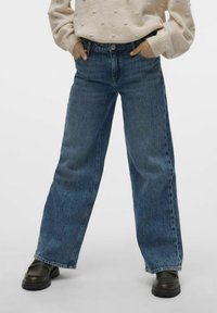 Personne debout les mains dans les poches, portant un jean large bleu, un pull beige texturé et des chaussures noires épaisses.