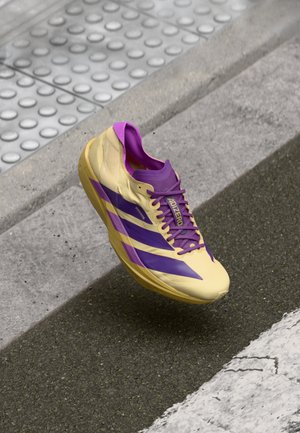 Geel en paars hardloopschoen met een gestroomlijnd ontwerp, voorzien van lichtgewicht materiaal, paarse veters en drie zijstrepen in paars.
