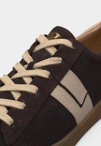 Σκούρο καφέ σουέντ αθλητικό παπούτσι με μπεζ κορδόνια και υφή λουστρίνι σε μπεζ χρώμα στη ρίγα στο πλάι, εμφανίζεται σε κοντινό πλάνο.