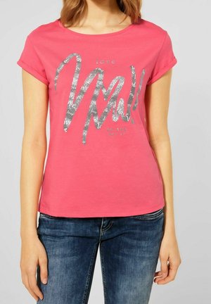 Kvinde iført en lyserød T-shirt med korte ærmer med sølvglimmertekst, der siger "love", og blå jeans, stående foran en ensfarvet baggrund.