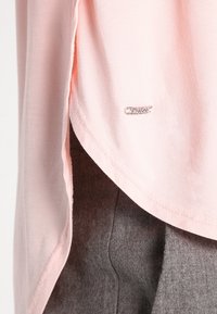 Hellrosa Stoff mit einem kleinen Metalletikett nahe der geschichteten Kante, drapiert über einem dunkelgrauen strukturierten Stoff darunter.