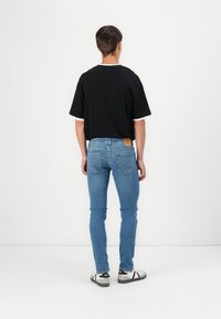 Černé tričko s krátkým rukávem a bílým lemem, modré slim-fit džíny a černo-bílé tenisky, viděno ze zadní strany. Dřevěná cedulka na džínách.