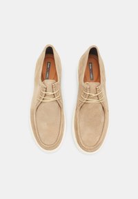 Pantofi din suede bej cu vârf rotund, șireturi decorative și talpă albă în contrast; textură netedă cu detalii de cusătură.