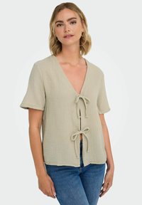 Lys beige struktureret top med V-udskæring, korte ærmer og to frontknuder, parret med denim jeans.