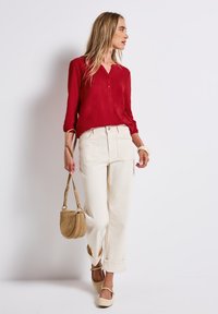 Donna con una blusa rossa, pantaloni bianchi arrotolati sui orli, scarpe beige e che porta una borsa a tracolla beige, in piedi contro uno sfondo bianco uniforme.