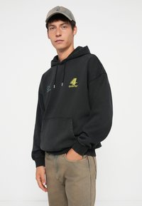 Hoodie noir avec poche avant, capuche à cordon de serrage, et texte graphique jaune. Porté avec un pantalon texturé brun clair. Le modèle se tient devant un fond neutre.