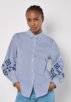Niebieska i biała koszula w paski z długimi, bufiastymi rękawami i haftowanymi detalami przy mankietach. Posiada kołnierz zapinany na guziki i guziki z przodu.