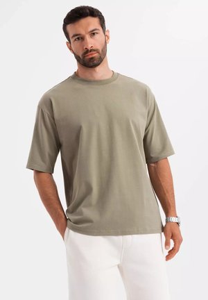 T-shirt a maniche corte in tessuto verde oliva, scollo rotondo, vestibilità rilassata, texture liscia. Abbinato a pantaloni bianchi e un orologio da polso.