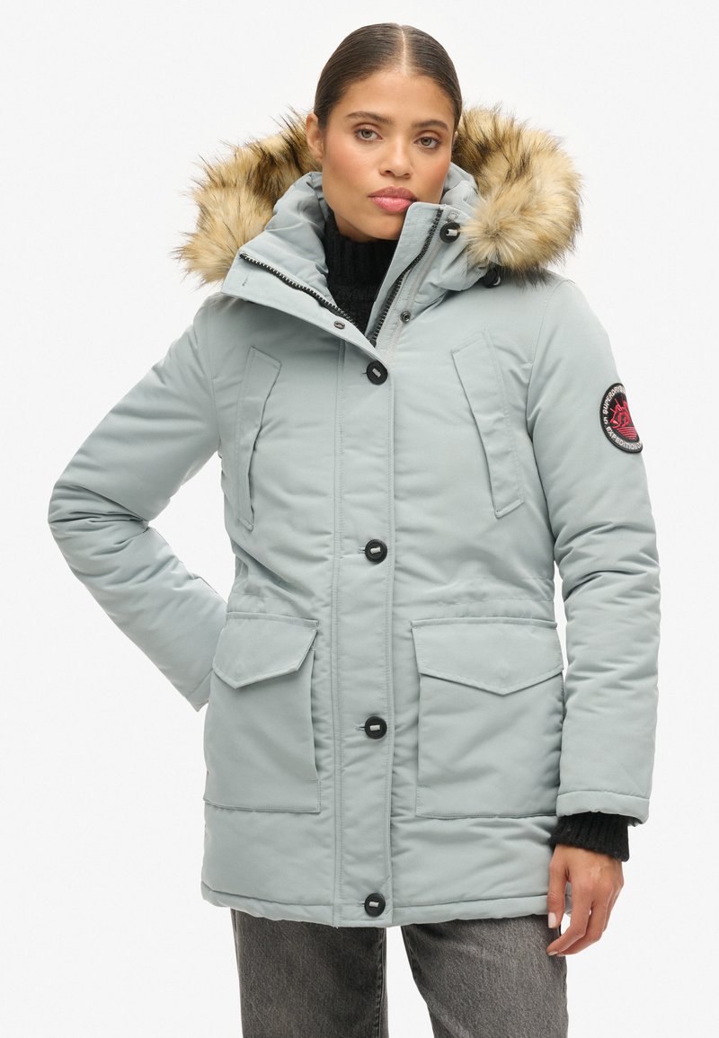 Parka leggera blu con cappuccio in pelliccia sintetica, chiusura con zip e bottoni, due grandi tasche frontali e un patch con logo sulla manica.
