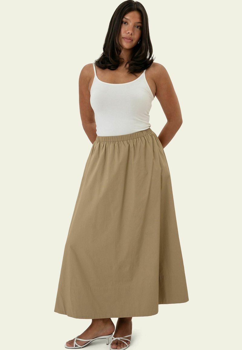 Femme portant un haut blanc sans manches, une longue jupe beige et des sandales blanches à talons, debout les mains dans le dos.