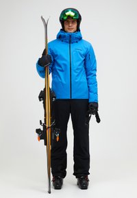 Blauwe waterdichte skijas met een capuchon, zwarte skipants, handschoenen en skischoenen; houdt ski's vast met oranje accenten en bindingen.