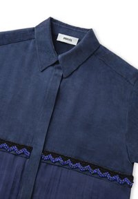 Camisa azul marino de tejido texturizado con cuello de punta. Presenta un adorno de cuentas en azul y negro a lo largo del pecho.