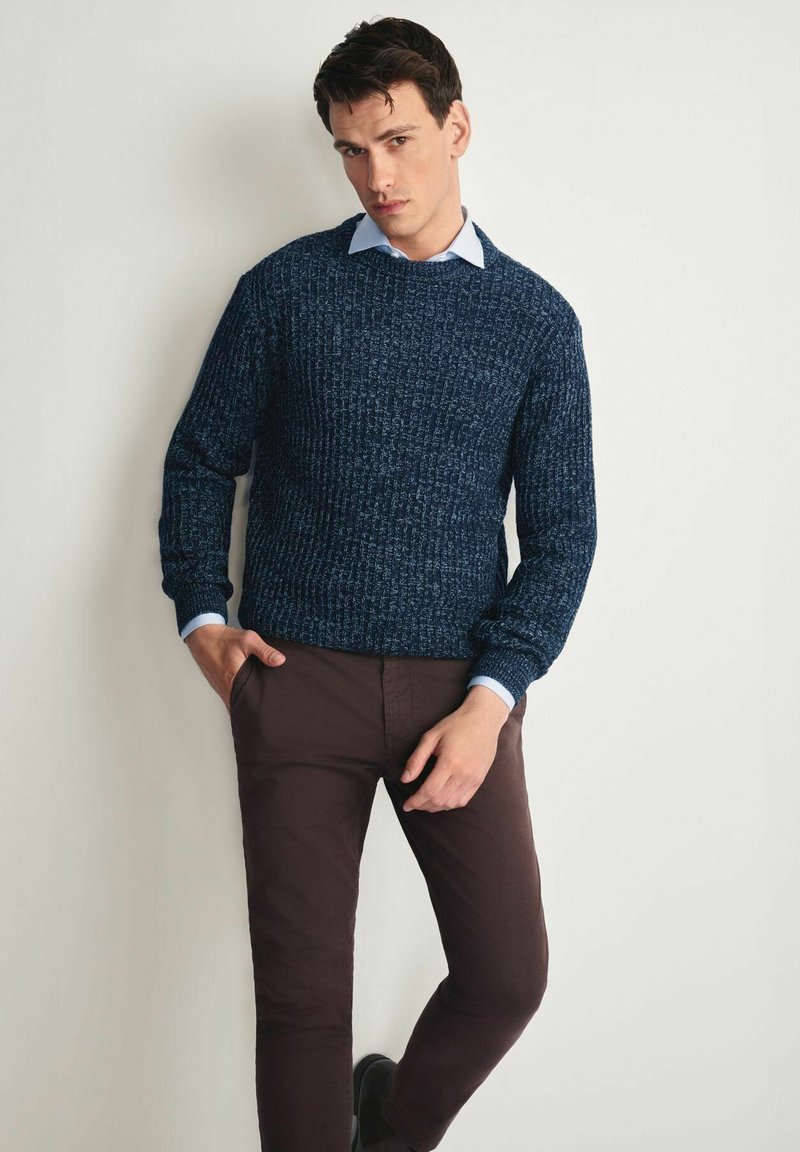 Granatowy sweter dzianinowy z teksturowanym wzorem, okrągłym dekoltem i długimi rękawami, noszony na jasnoniebieskiej koszuli z kołnierzykiem. Brązowe dopasowane spodnie.