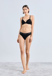 Must rinnahoidja õhukeste reguleeritavate rihmade ja sügava dekolteega, sobitatud mustade pükstega, millel on logo-ääris. Sile tekstuur.