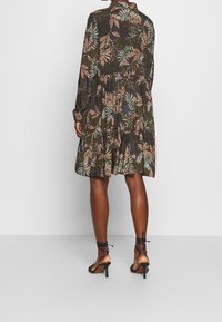 Langärmliges Kleid in Dunkelgrün mit Blumenmustern in Pink und Blau, ausgestattet mit einem gestuften Design und einer taillierten Taille. Kombiniert mit schwarzen Riemchen-Heels.