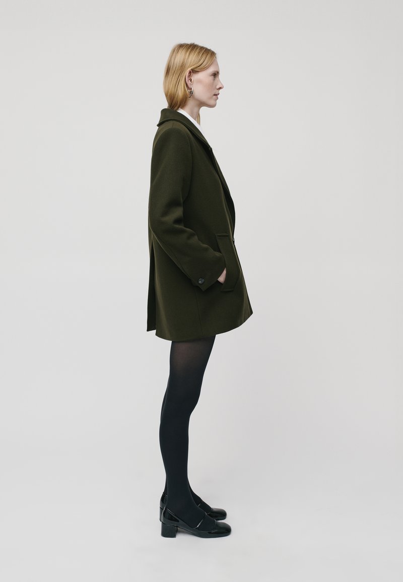 Manteau en mélange de laine vert olive avec col cranté, poches latérales et détails de boutons, porté avec des collants et des chaussures noirs, vu de profil.