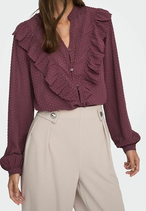 Donna che indossa una camicetta bordeaux a maniche lunghe con volant e puntini in rilievo, abbinata a pantaloni beige a vita alta con dettagli a bottoni.