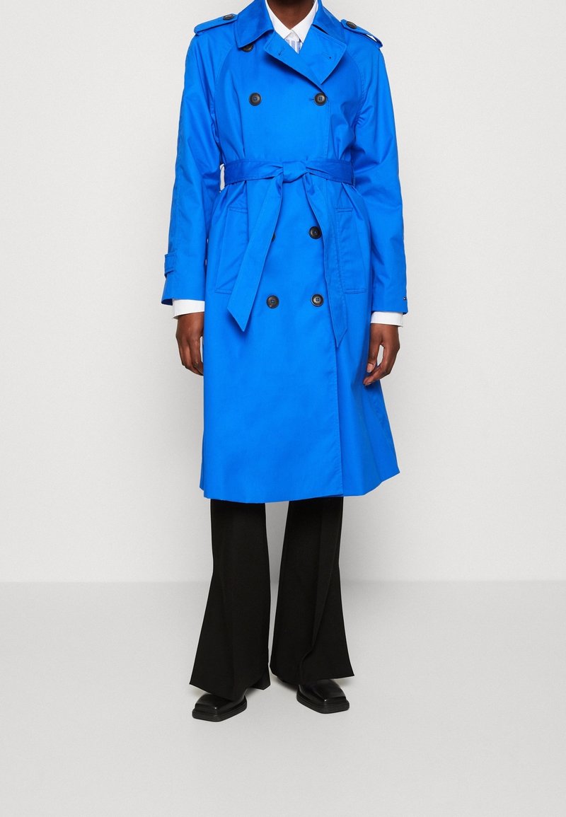 Niebieski płaszcz trencz z dwurzędowym zapięciem na guziki, paskiem w talii, naramiennikami i długimi rękawami, zestawiony z czarnymi rozszerzanymi spodniami.