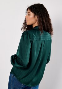 Blusa in satin verde con strisce verticali, maniche lunghe e colletto alla cinese. Presenta una vestibilità rilassata e dettagli sul pannello posteriore.