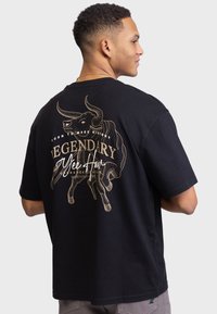 Mand iført sort t-shirt med stort guld okse-graphic og teksten "Legendary Yee Haw" på bagsiden, der står foran en ensfarvet lys baggrund.
