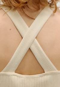 Beige, ribbelstof met gekruiste bandjes op de rug. De textuur is zacht en het ontwerp heeft een lage uitsnijding.