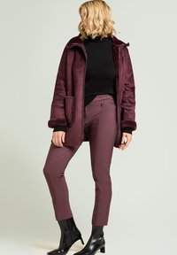 Abrigo de piel sintética burdeos con cuello y bolsillos laterales, combinado con pantalones burdeos ajustados. La modelo lleva botas negras de tacón bajo y un cuello alto negro.