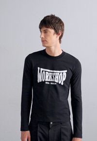 Svart langermet t-skjorte med hvit grafisk trykk som leser "WORKSHOP 032c BERLIN" på forsiden. Enkel rund hals og tettsittende design.