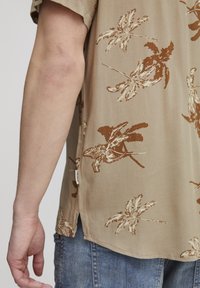 Camisa de manga corta en tejido beige con estampado floral en tonos marrones. Presenta una abertura lateral y una etiqueta en el dobladillo. Se lleva con vaqueros azules.