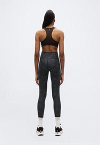 Γυναίκα που στέκεται πλάτη  με μαύρο αθλητικό σουτιέν από δίχτυ, ψηλόμεσο σκούρο γκρι κολάν, λευκές κάλτσες και μαύρα αθλητικά παπούτσια σε λευκό φόντο.