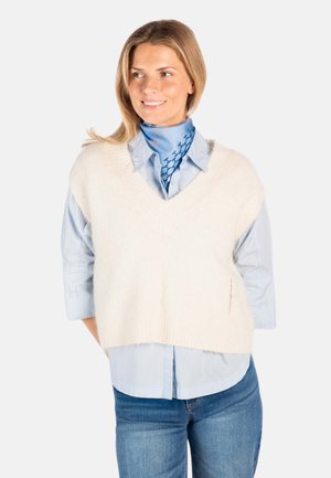 Kvinne iført lyseblå skjorte, hvit strikket vest uten ermer, blått mønstret hals tørkle og jeans, smilende og ser til siden.