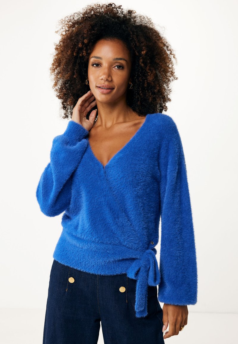 Blå fluffig omslagströja med V-ringning, långa ärmar och knytdetalj vid midjan. Mjuk textur, något lös passform, kombinerad med mörk denim.