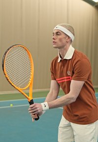 Teniso raketė su oranžine rankena ir baltais stygomis; žmogus rudame polo marškinėliuose su raudonomis juostelėmis, balduose šortuose ir baltu galvos juostu.