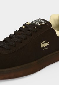Μαύρο αθλητικό παπούτσι Lacoste με μαύρα κορδόνια, κρεμ εσωτερικό και μικρό λευκό λογότυπο κροκοδείλου με "EST. 1933" στο πλάι.