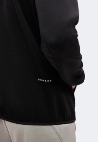 Μανίκι μαύρου μπουφάν Oakley και κάτω μέρος πλάτης, ορατό μπεζ παντελόνι, το χέρι του ατόμου εν μέρει μέσα στην τσέπη.