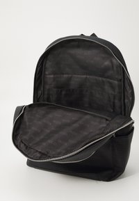 Sac à dos noir avec un extérieur texturé, une fermeture éclair argentée, et un intérieur présentant un motif répétitif du logo "GUESS" sur un tissu doux.