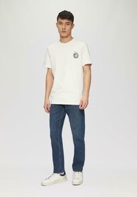 Witte t-shirt met een zwart grafisch ontwerp op de linkerborst, gecombineerd met donkerblauwe jeans en witte sneakers. Eenvoudig, casual ontwerp en pasvorm.