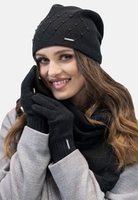 Zwarte gebreide beanie met diamanten patroon en kraalaccenten. Zwarte handschoenen en sjaal completeren de outfit, tentoongesteld tegen een neutrale achtergrond.