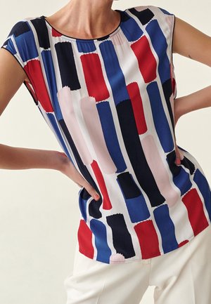 Top sans manches avec un motif abstrait en rectangles rouges, bleus, noirs et roses, porté avec un pantalon blanc, les mains sur les hanches.