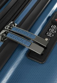 Dos tiradores metálicos de cremallera etiquetados como "REDOLZ" asegurados en un candado de combinación negro sobre una maleta azul texturizada.