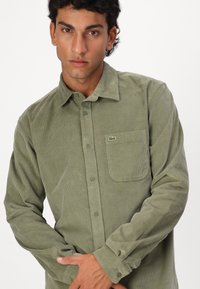 Olijfgroene corduroy overhemde met knoopsluiting aan de voorkant, lange mouwen, een enkele borstzak en geborduurd logo op de zak.