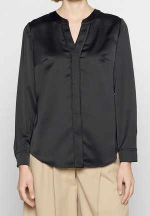 Czarna satynowa bluzka z długim rękawem, dekoltem w serek i ukrytymi guzikami z przodu, noszona z beżowymi plisowanymi spodniami.