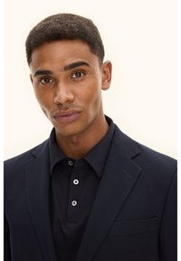 Marinblå kavaj med texturerad finish, bärs över en mörk pikétröja med tre knappar. Modellen har kort hår och jämn hudton.