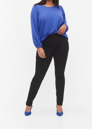 Mujer con blusa azul de manga larga, pantalones negros ajustados y zapatos de tacón azul, de pie sobre un fondo blanco.