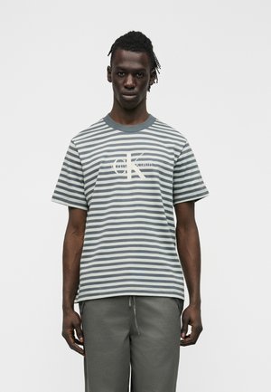 Mann mit kurzen Dreadlocks, der ein graues und weiß gestreiftes Calvin Klein T-Shirt und graue Hosen trägt, vor einem schlichten Hintergrund stehend.