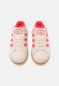 Jasnobeżowe zamszowe trampki z różowymi paskami, okrągłym noskiem i gumowymi podeszwami. Wnętrze wyściełane czerwonym materiałem z logo na języku. Zapinane na sznurowadła.