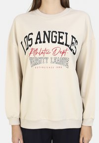 Felpa beige con scollo rotondo, design a spalla scesa e polsini a coste. Presenta testo stampato in nero e rosso: "LOS ANGELES Athletic Dept. VARSITY LEAGUE."
