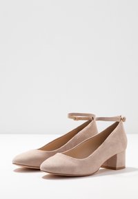 Scarpe beige in camoscio con tacco a blocco, punta rotonda e cinturino alla caviglia. Superficie texturizzata e design minimale. Tacco robusto e arrotondato.