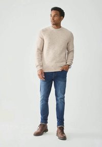 Homem com um suéter creme com manchas, jeans azuis e botas de couro castanho, em pé contra um fundo cinza claro simples.