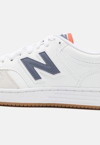 Bílé tenisky s šedým logem "N", semišovými akcenty a strukturovanou gumovou podrážkou. Mají kulatou špičku a ploché tkaničky pro klasický design.