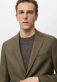 Mladić s maslinasto zelenim sakoom preko smeđeg pletenog džempera, gleda ravno ispred s neutralnim izrazom lica, na jednostavnoj svijetloj pozadini.